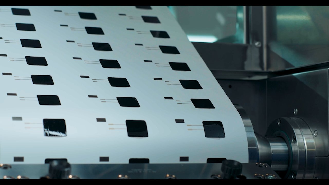 Sheet of black squares with thin lines on white material, moving through machinery.